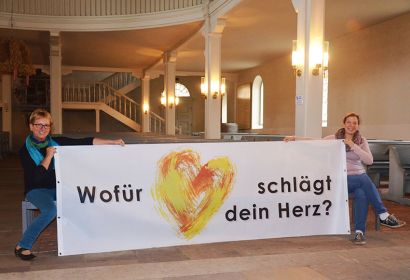 Mit einem Banner weisen Dörte Behn-Hartwig (links) und Pastorin Debora Becker auf die Ausstellung in der Brelinger Kirche hin. Foto: Andrea Hesse